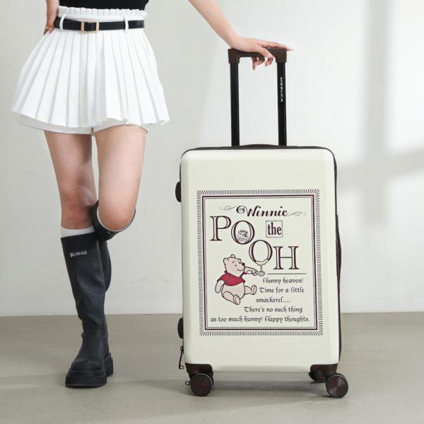 A young woman in a black shirt and white skirt, standing beside the Winnie the Pooh 20-inch suitcase, smiling while carrying a matching handbag.